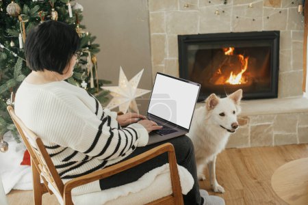 Téléchargez les photos : Heureuse femme âgée travaillant sur un ordinateur portable avec écran vide dans le salon de Noël élégant et festif avec un adorable chien blanc. Belle femme pigiste ayant télétravail ou achats en ligne - en image libre de droit
