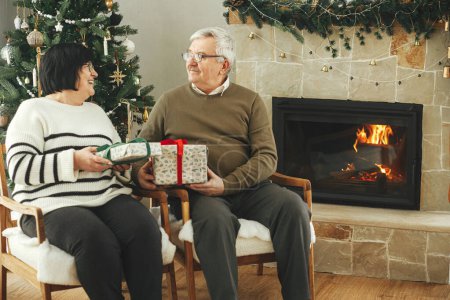 Foto de Hermosa pareja de ancianos que intercambian regalos de Navidad y se sientan en la chimenea caliente en la elegante sala de estar decorada festiva. Feliz familia mayor disfrutando de una noche acogedora con regalos. Felices fiestas - Imagen libre de derechos