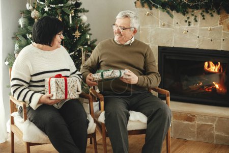 Téléchargez les photos : Beau couple de personnes âgées échangeant des cadeaux de Noël et assis à la cheminée chaude dans le salon décoré élégant et festif. Heureuse famille aînée profitant d'une soirée confortable avec des cadeaux. Joyeuses fêtes - en image libre de droit