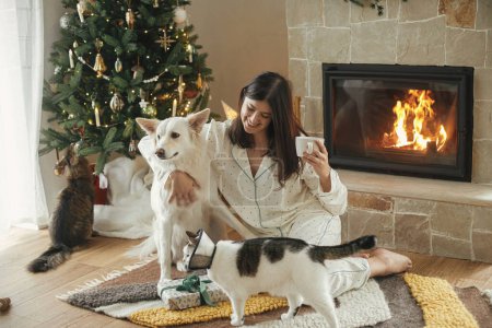 Téléchargez les photos : Belle femme en pyjama élégant avec thé chaud relaxant avec chien et chat à la cheminée confortable, profiter du matin de Noël dans le salon décoré festif. Joyeux Noël ! Vacances d'hiver avec animaux domestiques - en image libre de droit