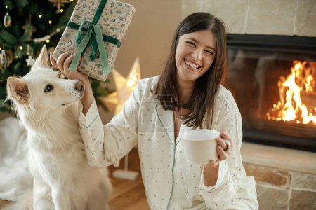 Téléchargez les photos : Femme heureuse en pyjama élégant avec thé chaud relaxant avec chien mignon à la cheminée confortable, profiter du matin de Noël dans le salon décoré de fêtes avec des cadeaux. Joyeux Noël ! Vacances d'hiver - en image libre de droit