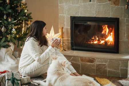 Téléchargez les photos : Confortable matin de Noël. Belle femme en pyjama élégant tenant le thé chaud et relaxant avec chien mignon à la cheminée dans le salon décoré festif. Joyeux Noël ! Vacances d'hiver avec animaux domestiques - en image libre de droit