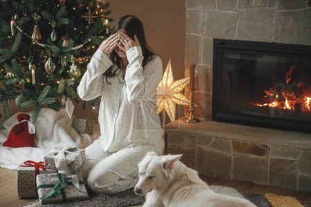 Téléchargez les photos : Surprise du matin de Noël. Belle femme en pyjama et mignon chien blanc fermant les yeux avant les cadeaux de Noël élégants à l'arbre décoré dans le salon de fête. Joyeux Noël! - en image libre de droit