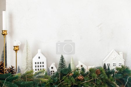 Téléchargez les photos : Élégante cheminée décorée de Noël. Arbres de Noël modernes, maisons, cônes de pin et branches d'épinette sur cheminée manteau avec cloches en laiton et ruban. Décor scandinave de salon de Noël - en image libre de droit