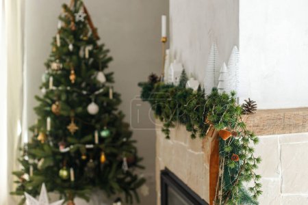 Foto de Elegante chimenea de Navidad decorada. Modernos árboles de Navidad, casas, conos de pino y ramas de abeto en la chimenea mantel con campanas de latón y cinta. Navidad escandinava decoración de la sala de estar - Imagen libre de derechos