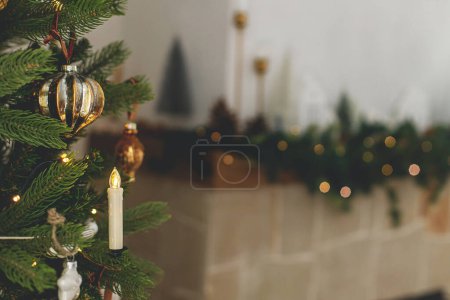 Photo for Stylish decorated christmas tree with vintage golden baubles and candles against fireplace mantel with lights. Atmospheric winter holidays. Merry christmas! - Royalty Free Image