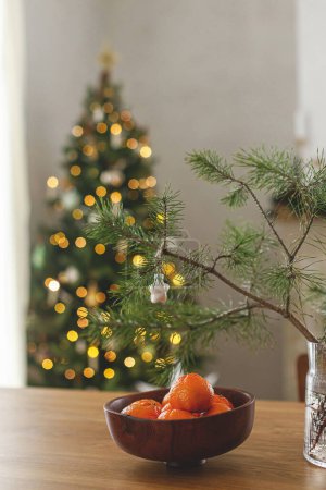 Téléchargez les photos : Mandarines fraîches dans un bol en bois et branche d'épinette avec boule étoilée contre élégant arbre de Noël décoré avec des lumières dorées. Vacances d'hiver atmosphériques à la maison. Joyeux Noël! - en image libre de droit