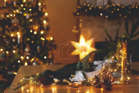 Foto de Vela con estilo, luces doradas, conos de pino y adornos en la mesa de madera contra el elegante árbol de Navidad decorado y la chimenea con iluminación festiva. Vacaciones de invierno atmosféricas en el hogar festivo - Imagen libre de derechos