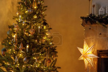 Foto de ¡Feliz Navidad! Nochebuena atmosférica. Elegante estrella iluminada de Navidad, árbol de Navidad decorado con luces doradas y decoración festiva en la chimenea mantel en la habitación escandinava. - Imagen libre de derechos