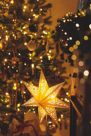 Photo for Atmospheric christmas eve. Stylish christmas illuminated star, christmas tree with golden lights and festive decorated fireplace in evening scandinavian room. Merry Christmas! - Royalty Free Image