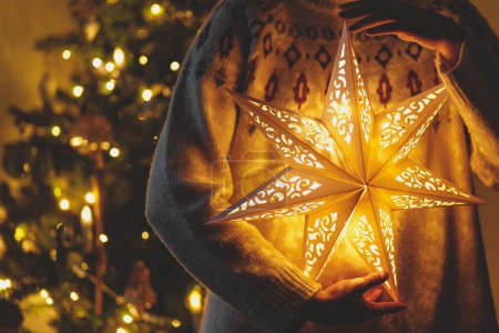 Photo for Merry Christmas! Stylish illuminated golden star in hands on background of modern decorated christmas tree with golden lights in evening room. Atmospheric magical christmas eve - Royalty Free Image