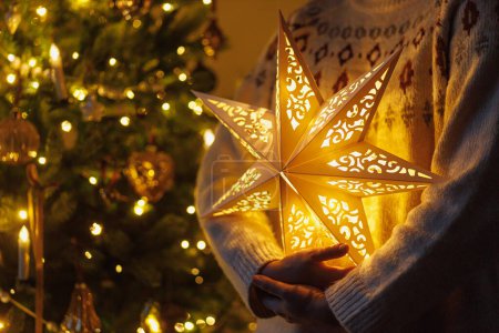 Téléchargez les photos : Joyeux Noël ! Élégante étoile dorée illuminée dans les mains sur fond d'arbre de Noël moderne décoré avec des lumières dorées dans la salle du soir. Atmosphérique magique veille de Noël - en image libre de droit