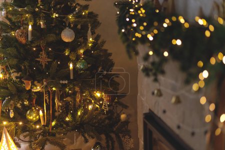 Photo for Stylish illuminated christmas tree with vintage baubles and festive decorated fireplace with golden lights in evening scandinavian room. Atmospheric christmas eve. Merry Christmas! - Royalty Free Image