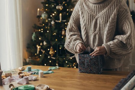 Téléchargez les photos : Femme en pull confortable enveloppant cadeau de Noël élégant en papier d'emballage festif avec des rubans, des ornements vintage, des arcs sur une table en bois. Vacances d'hiver dans l'atmosphère, emballage cadeau de Noël - en image libre de droit