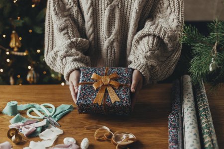 Téléchargez les photos : Joyeux Noël ! Femme en pull confortable tenant cadeau de Noël élégant en papier d'emballage festif avec arc, ruban, ornements vintage sur table en bois. Vacances d'hiver dans l'atmosphère - en image libre de droit