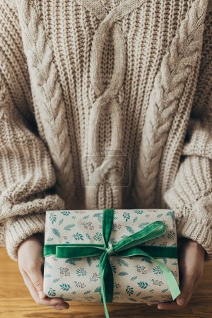 Foto de ¡Feliz Navidad! Mujer en suéter acogedor celebración elegante regalo de Navidad en papel de regalo festivo con lazo, cinta en la mesa de madera. Vacaciones de invierno atmosféricas - Imagen libre de derechos