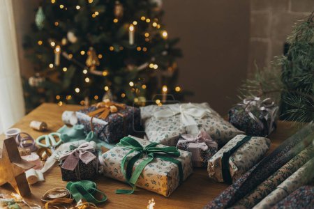 Téléchargez les photos : Joyeux Noël ! Cadeau de Noël élégant, papier d'emballage festif, rubans, ciseaux, ornements vintage, cloches en laiton sur une table en bois sur fond de lumières atmosphériques de l'arbre de Noël. - en image libre de droit
