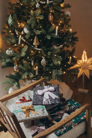 Foto de ¡Feliz Navidad! Elegantes regalos de Navidad envueltos en papel festivo con arcos, adorno vintage y juguete en silla moderna en el fondo de las luces del árbol de Navidad. Vacaciones de invierno atmosféricas - Imagen libre de derechos