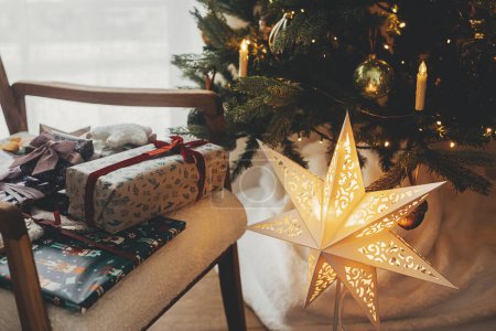 Téléchargez les photos : Joyeux Noël ! Cadeaux de Noël élégants enveloppés dans du papier de fête avec des arcs, des jouets vintage sur une chaise moderne sur fond de lumières d'arbre de Noël et d'étoile dorée. Vacances d'hiver dans l'atmosphère - en image libre de droit
