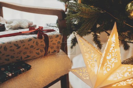 Foto de ¡Feliz Navidad! Elegantes regalos de Navidad envueltos en papel festivo con arcos, juguetes vintage en silla moderna en el fondo de las luces del árbol de Navidad y la estrella dorada. Vacaciones de invierno atmosféricas - Imagen libre de derechos
