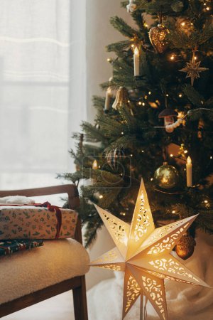 Foto de ¡Feliz Navidad! Elegantes regalos de Navidad envueltos en papel festivo con arcos, juguetes vintage en silla moderna en el fondo de las luces del árbol de Navidad y la estrella dorada. Vacaciones de invierno atmosféricas - Imagen libre de derechos