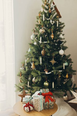 Foto de Elegantes regalos de navidad en papel de regalo festivo con arcos y adornos vintage sobre mesa de madera sobre fondo de luces de árboles de navidad dorados. ¡Felices fiestas! Nochebuena atmosférica - Imagen libre de derechos