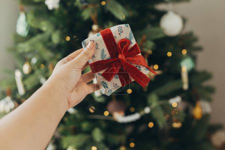 Téléchargez les photos : Main tenant cadeau de Noël élégant en papier d'emballage festif avec arc sur fond de lumières de sapin de Noël. Joyeuses fêtes ! Temps de Noël atmosphérique - en image libre de droit