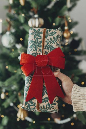 Foto de Mano en suéter acogedor celebración elegante regalo de Navidad en papel de regalo festivo con lazo en el fondo de las luces del árbol de Navidad. ¡Felices fiestas! Tiempo de Navidad atmosférica - Imagen libre de derechos