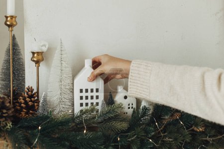 Photo for Decorating christmas fireplace with lights, fir branches, white little houses decoration. Hands in cozy sweater hanging decoration on mantel in modern farmhouse. Atmospheric winter holiday - Royalty Free Image