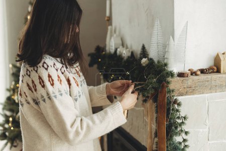 Téléchargez les photos : Femme en pull confortable accrochant une décoration élégante sur le manteau dans une ferme moderne. Décorer cheminée de Noël avec ruban de velours et cloches vintage sur branches de sapin. Vacances d'hiver dans l'atmosphère - en image libre de droit
