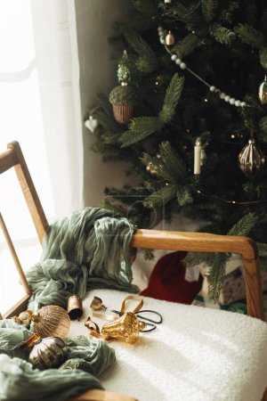 Téléchargez les photos : Élégantes boules vintage de Noël, rubans, cloches en laiton et ciseaux sur une chaise blanche confortable sous l'arbre. Ornements de Noël esthétique. Décorer l'arbre de Noël. Joyeux Noël! - en image libre de droit