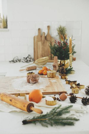 Téléchargez les photos : Pâte à pain d'épice avec farine sur planche de bois, rouleau à pâtisserie, découpeuses en métal doré, épices de cuisson et décorations festives sur le comptoir dans la cuisine blanche moderne. Faire des biscuits au pain d'épice de Noël - en image libre de droit