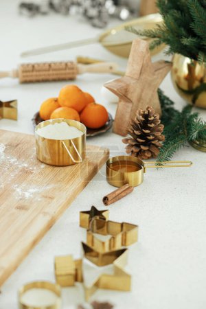 Téléchargez les photos : Faire des biscuits de pain d'épice de Noël dans la cuisine blanche moderne. Pâte à pain d'épice sur carton en bois, farine, rouleau à pâtisserie, coupe-métal doré, épices de cuisson et décorations festives sur le comptoir - en image libre de droit