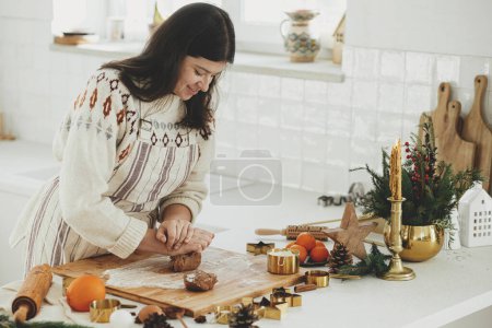 Téléchargez les photos : Femme faisant des biscuits de pain d'épice de Noël dans la cuisine blanche moderne. Pâte à pain d'épice à pétrir à la main sur une planche en bois avec farine, rouleau à pâtisserie, coupe-métal doré, épices de cuisson sur le comptoir - en image libre de droit