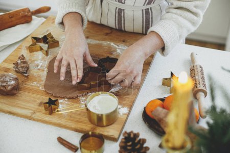 Photo for Hands cutting gingerbread dough with festive golden metal cutters on wooden board with flour, cooking spices, festive decorations in modern white kitchen. Woman making christmas gingerbread cookies - Royalty Free Image