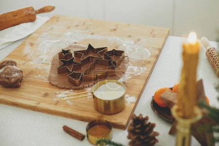 Foto de Hacer galletas de jengibre de Navidad. Masa de pan de jengibre con cortadores de metal dorado festivos en tablero de madera con harina, especias de cocina, decoraciones festivas en la cocina blanca moderna - Imagen libre de derechos