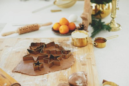 Téléchargez les photos : Pâte à pain d'épice avec découpeuses métalliques dorées festives sur planche de bois avec farine, épices de cuisson, décorations festives dans la cuisine blanche moderne. Faire des biscuits au pain d'épice de Noël - en image libre de droit