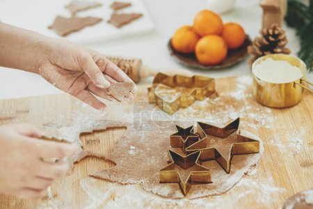 Foto de Mujer haciendo galletas de jengibre de Navidad se cierran en la cocina blanca moderna. Manos cortando masa de jengibre con cortadores festivos de metal dorado con especias y decoraciones para cocinar - Imagen libre de derechos