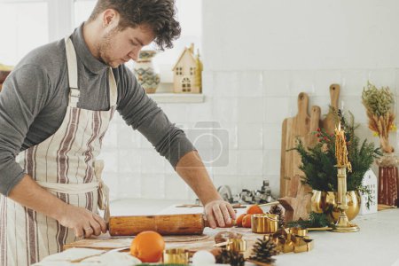 Foto de Hombre haciendo galletas de jengibre de Navidad en la cocina blanca moderna. Mano amasando masa de jengibre en tablero de madera con harina, rodillo, cortadores de metal dorado, especias de cocina en la encimera - Imagen libre de derechos