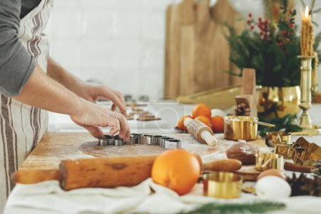 Téléchargez les photos : Homme faisant des biscuits de pain d'épice de Noël fermer dans la cuisine blanche moderne. Mains coupant la pâte de pain d'épice avec des couteaux métalliques dorés festives avec des épices et des décorations de cuisson - en image libre de droit