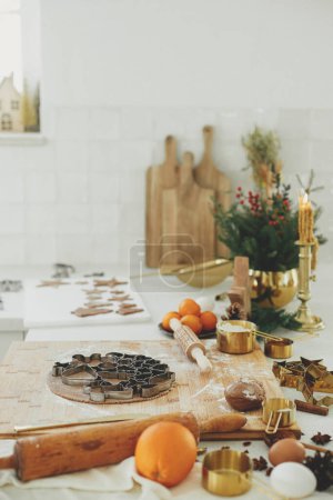 Foto de Hacer galletas de jengibre de Navidad. Masa de pan de jengibre con cortadores de metal festivos en tablero de madera con harina, especias de cocina, decoraciones festivas en la cocina blanca moderna - Imagen libre de derechos
