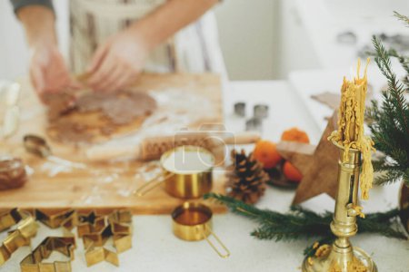 Foto de Vela vintage en el fondo del hombre haciendo galletas de jengibre de Navidad en la cocina blanca moderna. Manos cortando masa de jengibre con cortadores de metal dorado festivos en el mostrador con decoraciones - Imagen libre de derechos