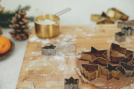 Foto de Masa de pan de jengibre con cortadores de metal dorado festivos en tablero de madera con harina, especias de cocina, decoraciones festivas en la cocina blanca moderna. Hacer galletas de jengibre de Navidad - Imagen libre de derechos