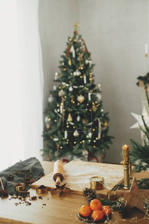 Foto de Harina sobre tabla de madera, rodillo, cortadores de metal dorado, especias de cocina y decoraciones festivas en mesa rústica contra el elegante árbol de Navidad decorado. Hacer galletas de jengibre de Navidad - Imagen libre de derechos