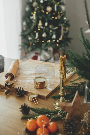 Téléchargez les photos : Farine sur panneau de bois, rouleau à pâtisserie, coupe-métal doré, épices de cuisine et décorations festives sur table rustique contre l'arbre de Noël décoré élégant. Faire des biscuits au pain d'épice de Noël - en image libre de droit