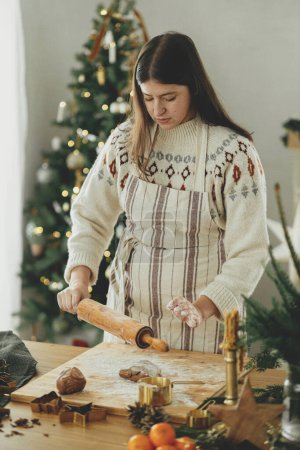 Photo for Woman making christmas gingerbread cookies. Hands kneading gingerbread dough with rolling pin, golden cutters, cooking spices, festive decorations on rustic table against stylish christmas tree - Royalty Free Image