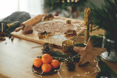 Foto de Masa de pan de jengibre con cortadores de oro, rodillo, especias de cocina y decoraciones festivas en la mesa de madera contra el elegante árbol de Navidad. Hacer galletas de jengibre de Navidad, tiempo de vacaciones - Imagen libre de derechos