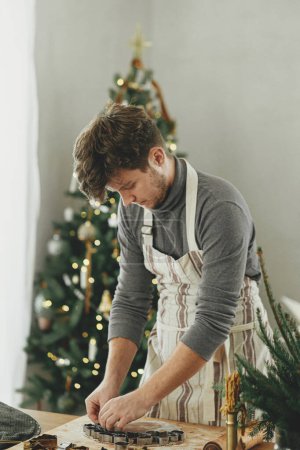 Photo for Hands cutting gingerbread dough with festive cutters on rustic table with holiday decorations against stylish christmas tree. Man making christmas gingerbread cookies, atmospheric holiday time - Royalty Free Image