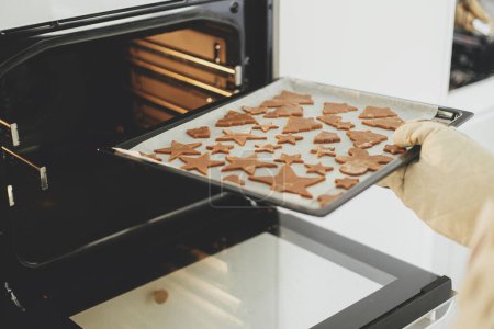 Foto de Mano en manoplas de horno poniendo bandeja con galletas de jengibre en formas navideñas festivas en horno moderno en cocina blanca. Hacer galletas de jengibre de Navidad - Imagen libre de derechos