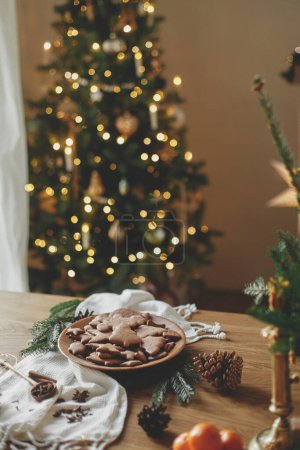 Téléchargez les photos : Délicieux biscuits au pain d'épice dans une assiette en bois sur une table rustique contre les lumières élégantes de l'arbre de Noël bokeh. Biscuits de pain d'épice de Noël frais cuits au four, temps des fêtes atmosphériques - en image libre de droit